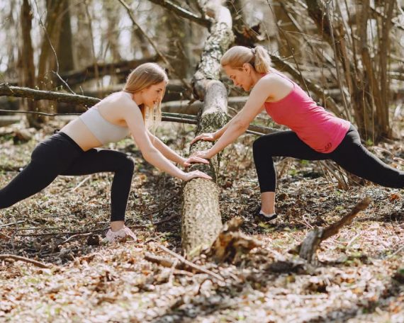 Side view of young female athletes in activewear stretching legs together using fallen tree during fitness workout in spring forest adhering to healthy lifestyle